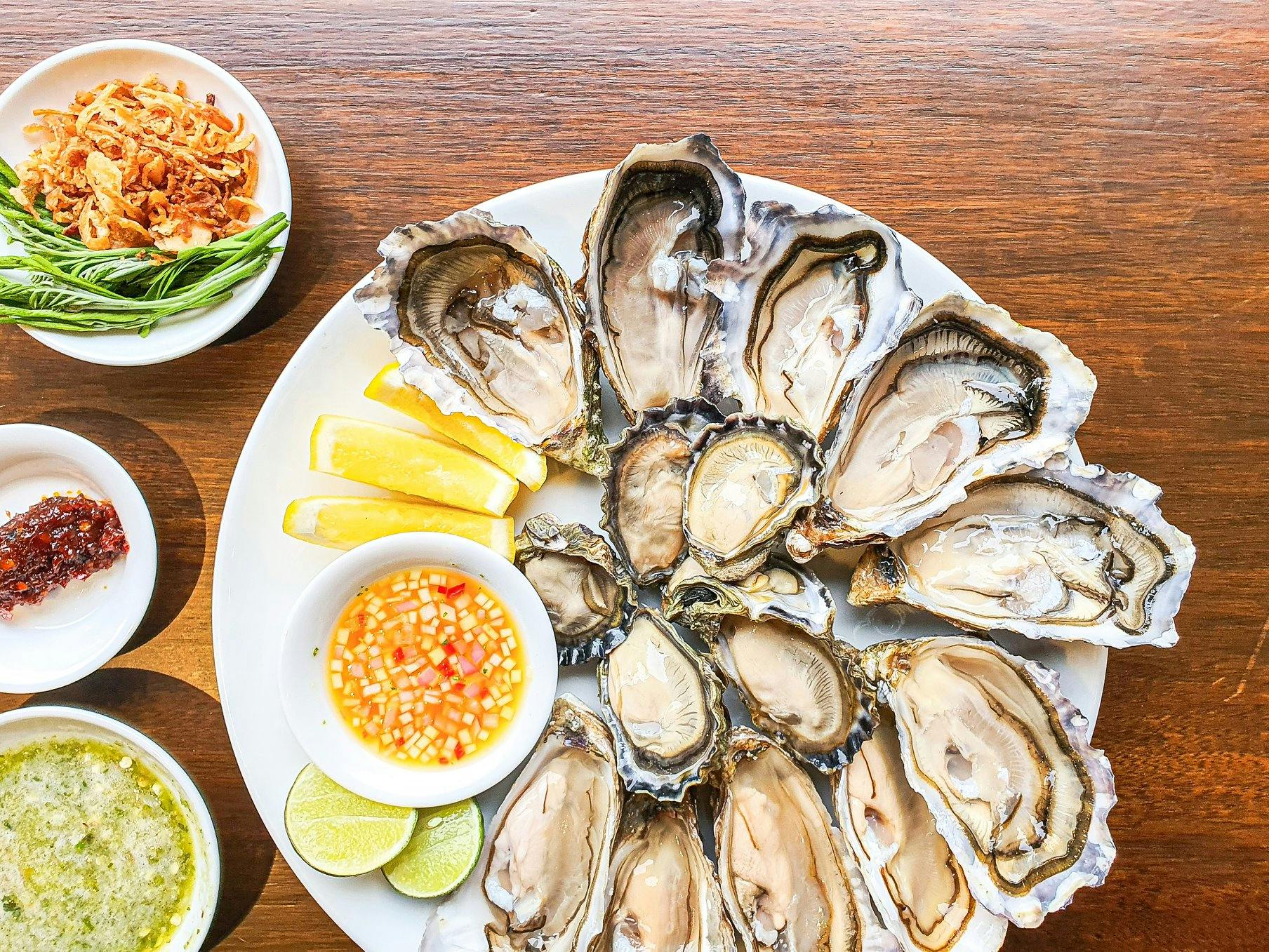 A plate of opened oysters on a wooden table, accompanied by lemon slices, lime wedges, and dipping sauces.