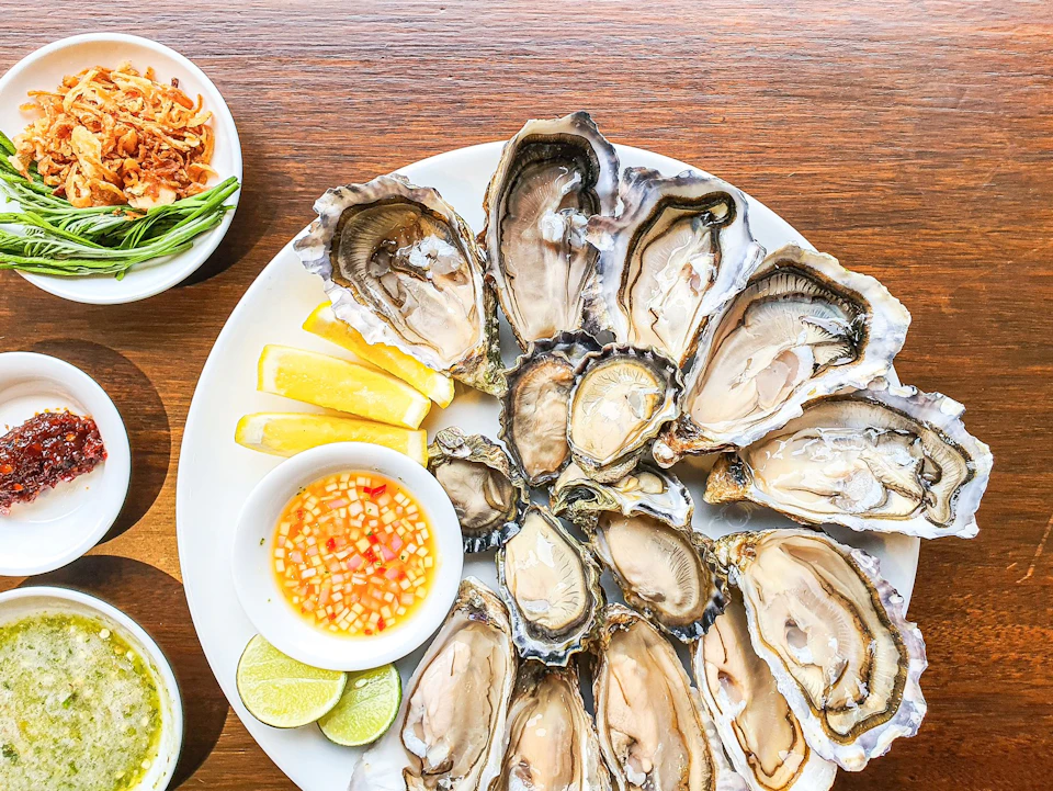 A plate of opened oysters on a wooden table, accompanied by lemon slices, lime wedges, and dipping sauces.