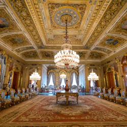 Utsmyckat rum med detaljerade tak, kristallkronor, blå klädda stolar klädda mot väggar och stora fönster med draperier.