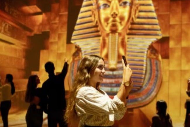 A woman takes a photo with her phone in front of a large illuminated display of a golden pharaoh mask in a museum-like setting.
