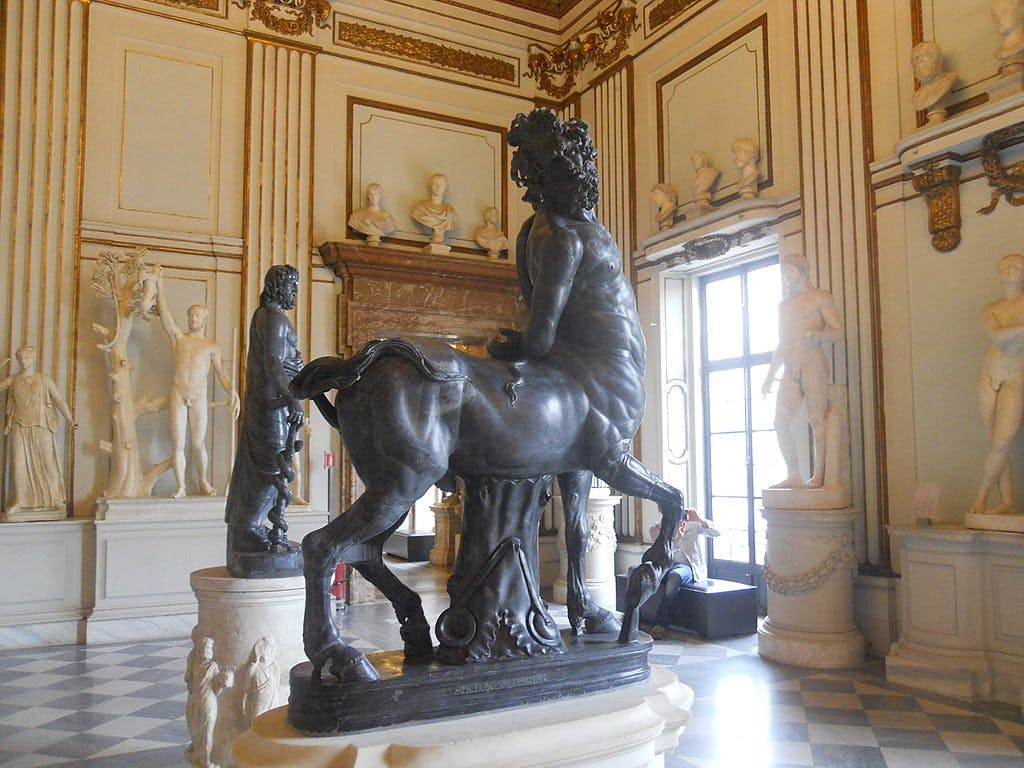 Sculpture of a centaur in an ornate room filled with classical statues and large windows, featuring decorative moldings and tiled flooring.