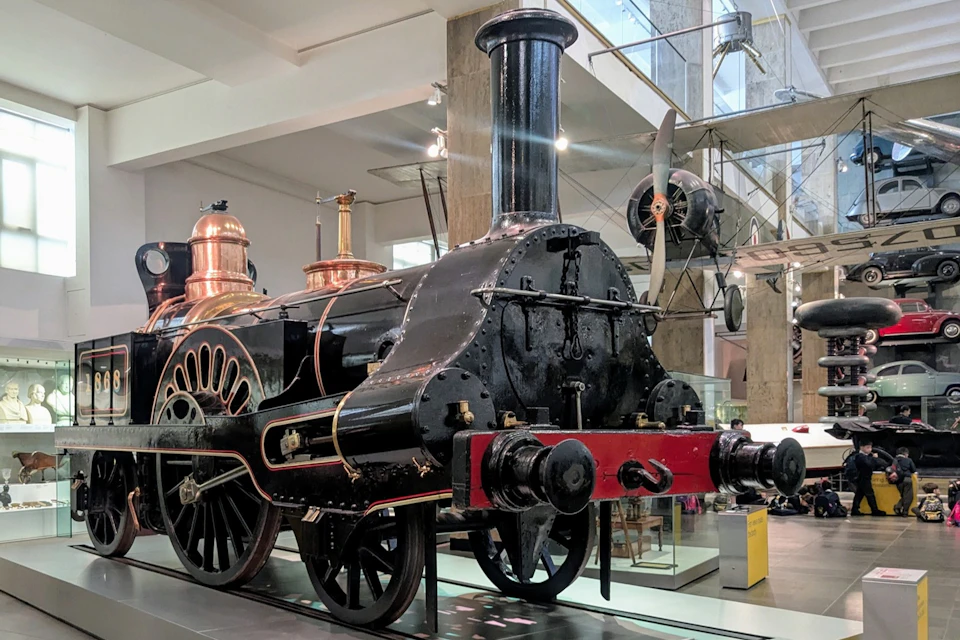 A historic steam locomotive displayed indoors, featuring black and red colors with copper details, in a museum setting.