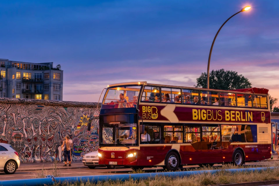 A red double-decker tour bus on a street near a graffiti-covered wall at sunset, with people visible on and around the bus.