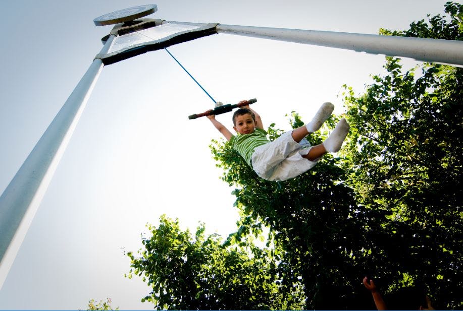 Barn i en randig skjorta svänger högt på en dragkedja, med träd och en klar himmel i bakgrunden.