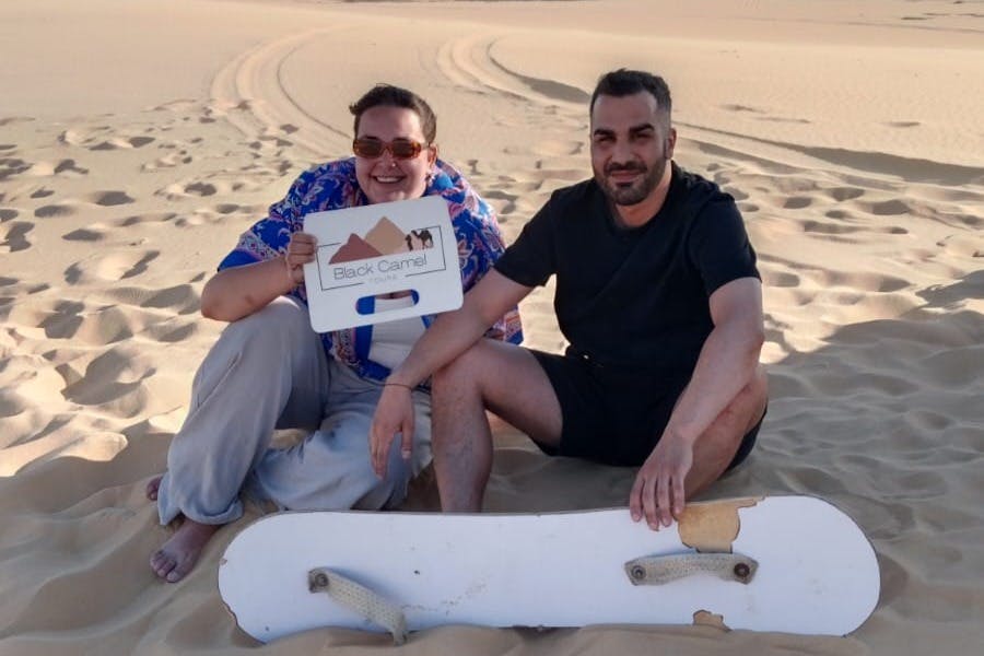 Two people sitting in the sand, one holding a "Black Camel" sign, with a sandboard in front of them and sand dunes in the background.