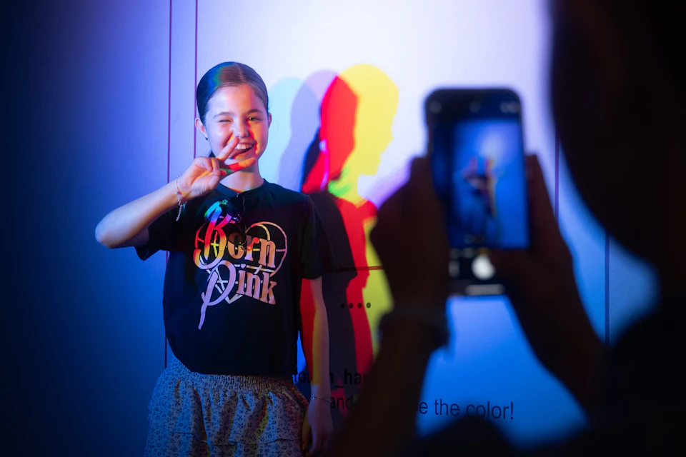 A person wearing a "Born Pink" shirt poses with a peace sign, illuminated by colorful lighting, creating shadow effects on the wall.