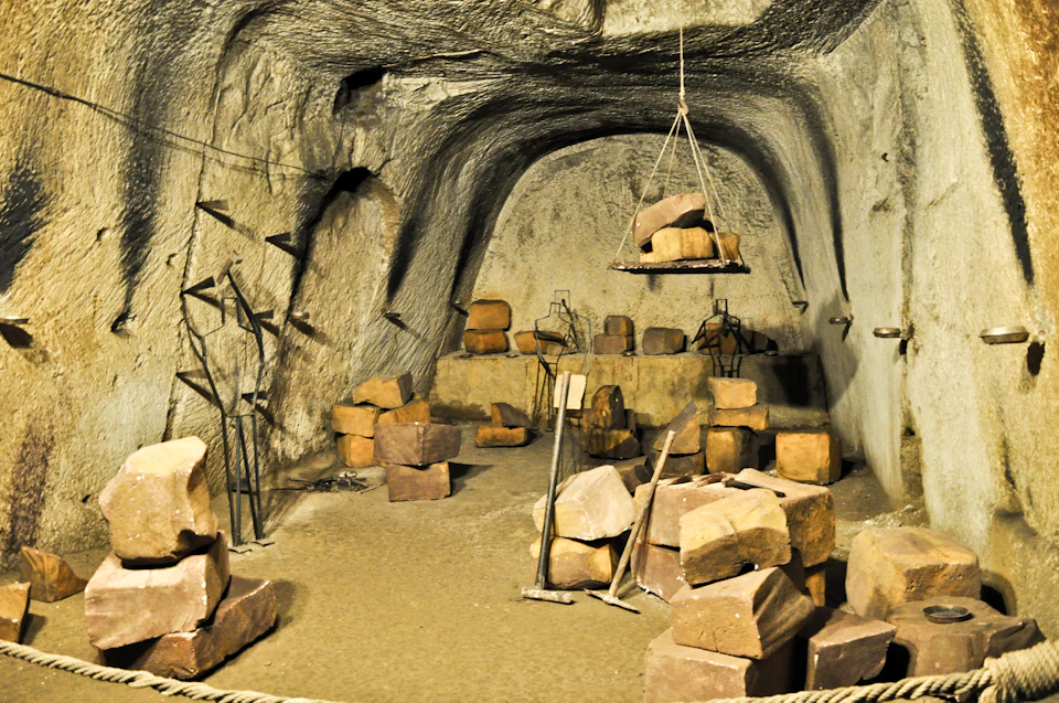 Underground cave with large stone blocks, metal tools, and a pulley system. Various construction and support structures are visible.