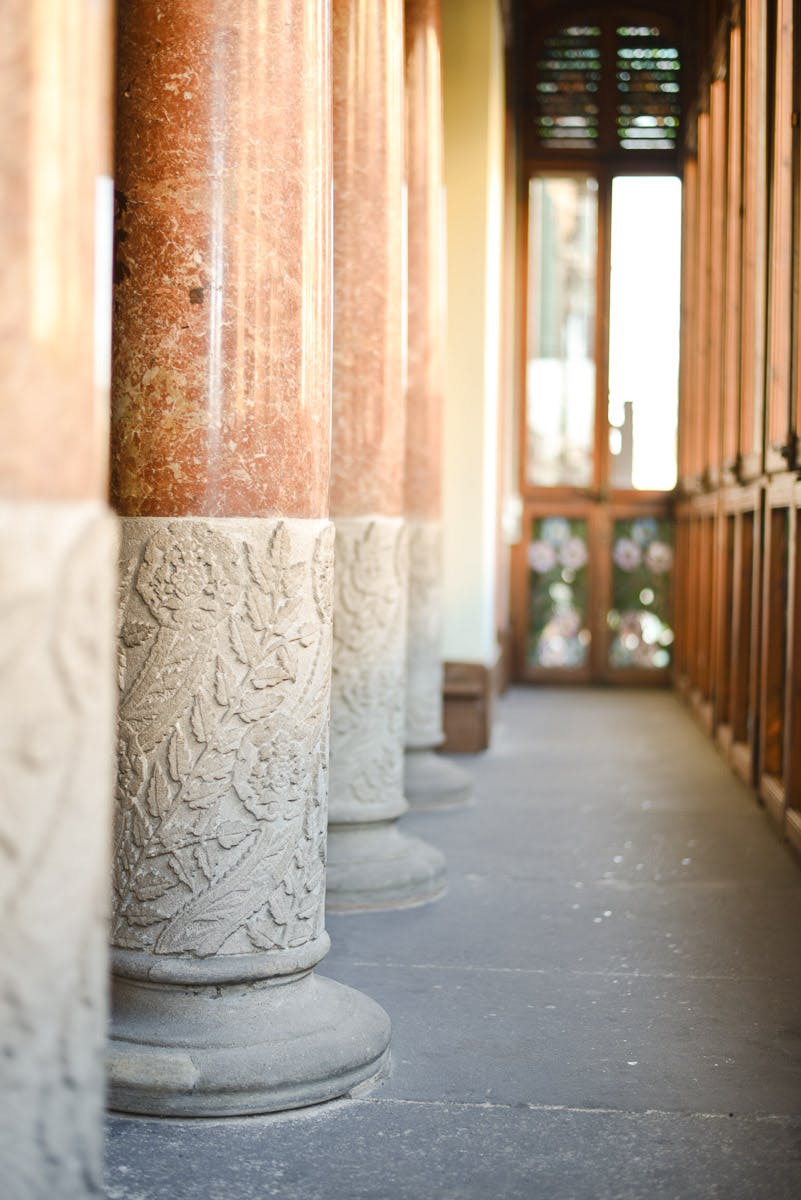 Gros plan sur des colonnes décoratives en pierre avec des sculptures florales ornées, alignant un couloir avec des portes en bois vitrées à l'arrière-plan.