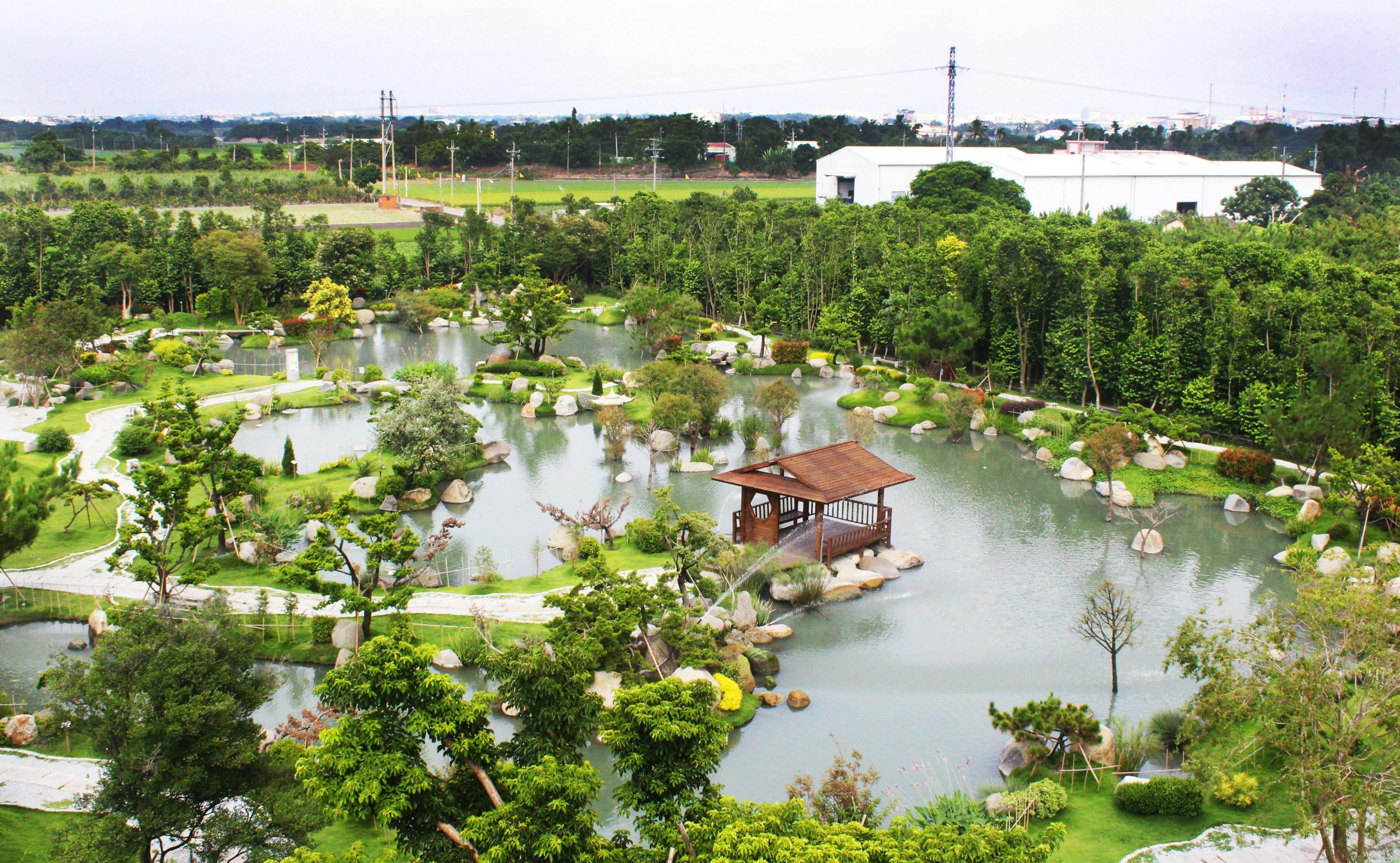 A serene garden with a pond, trees, rocks, and a wooden pavilion surrounded by greenery on a cloudy day.