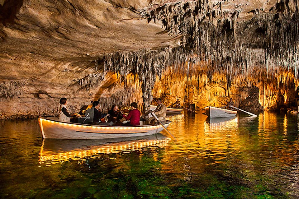 People in boats glide through an illuminated cave with stalactites, reflected in the water.