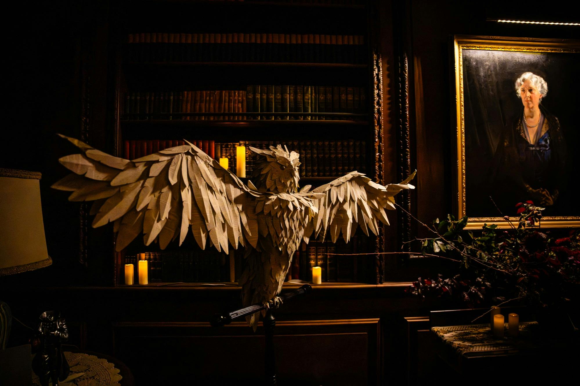 Ένα μεγάλο ξύλινο άγαλμα κουκουβάγιας με απλωμένα φτερά εκτίθεται σε μια αμυδρά φωτισμένη βιβλιοθήκη, που περιβάλλεται από ράφια και κεριά.