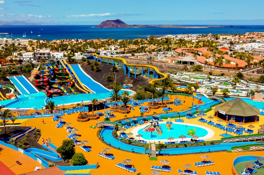 A colorful water park with pools, slides, and lounging areas, set against a coastal town and ocean backdrop.