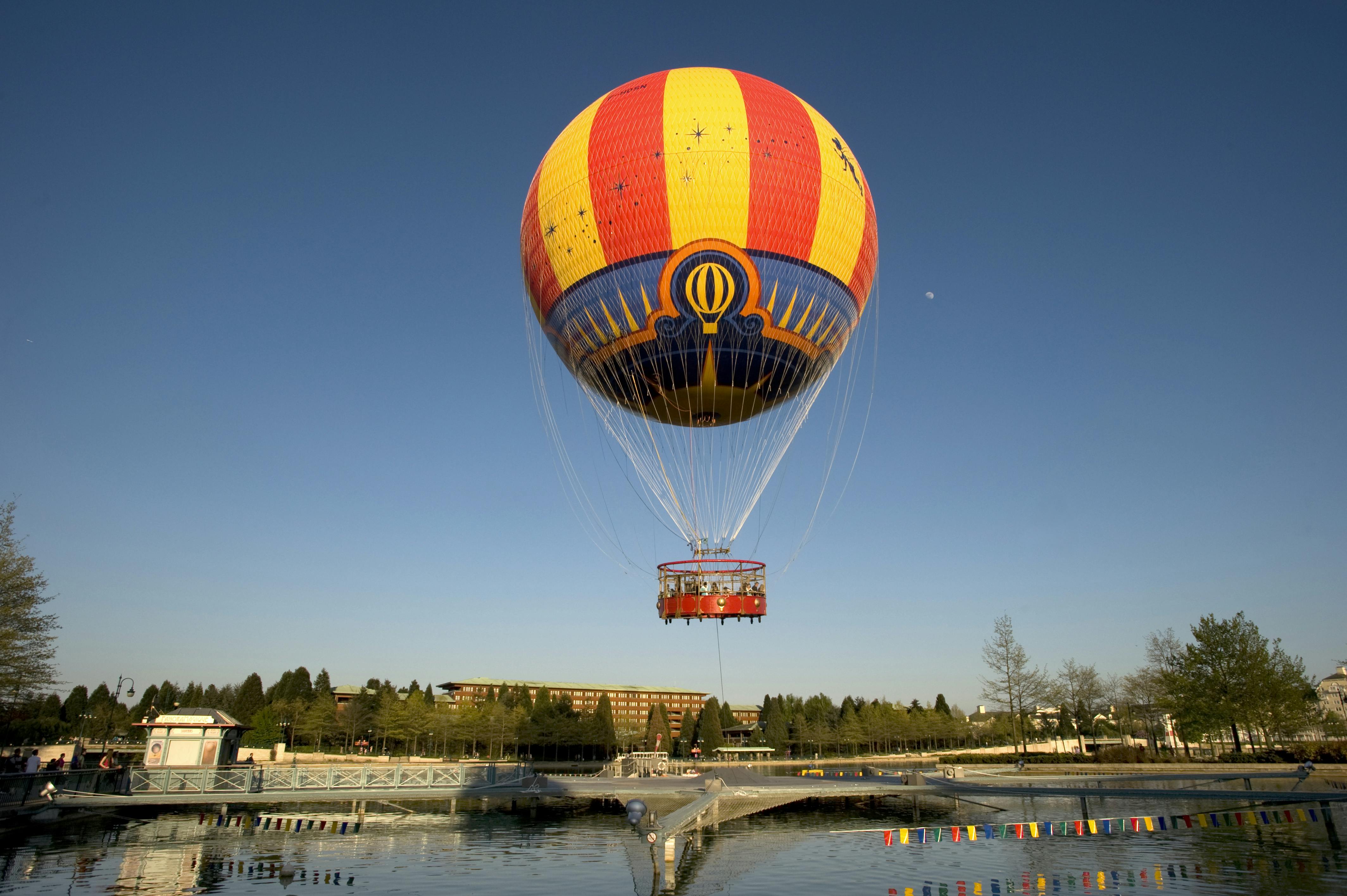 Une montgolfière colorée est attachée au-dessus d'un étang avec un quai, entouré d'arbres et de bâtiments sous un ciel bleu clair.