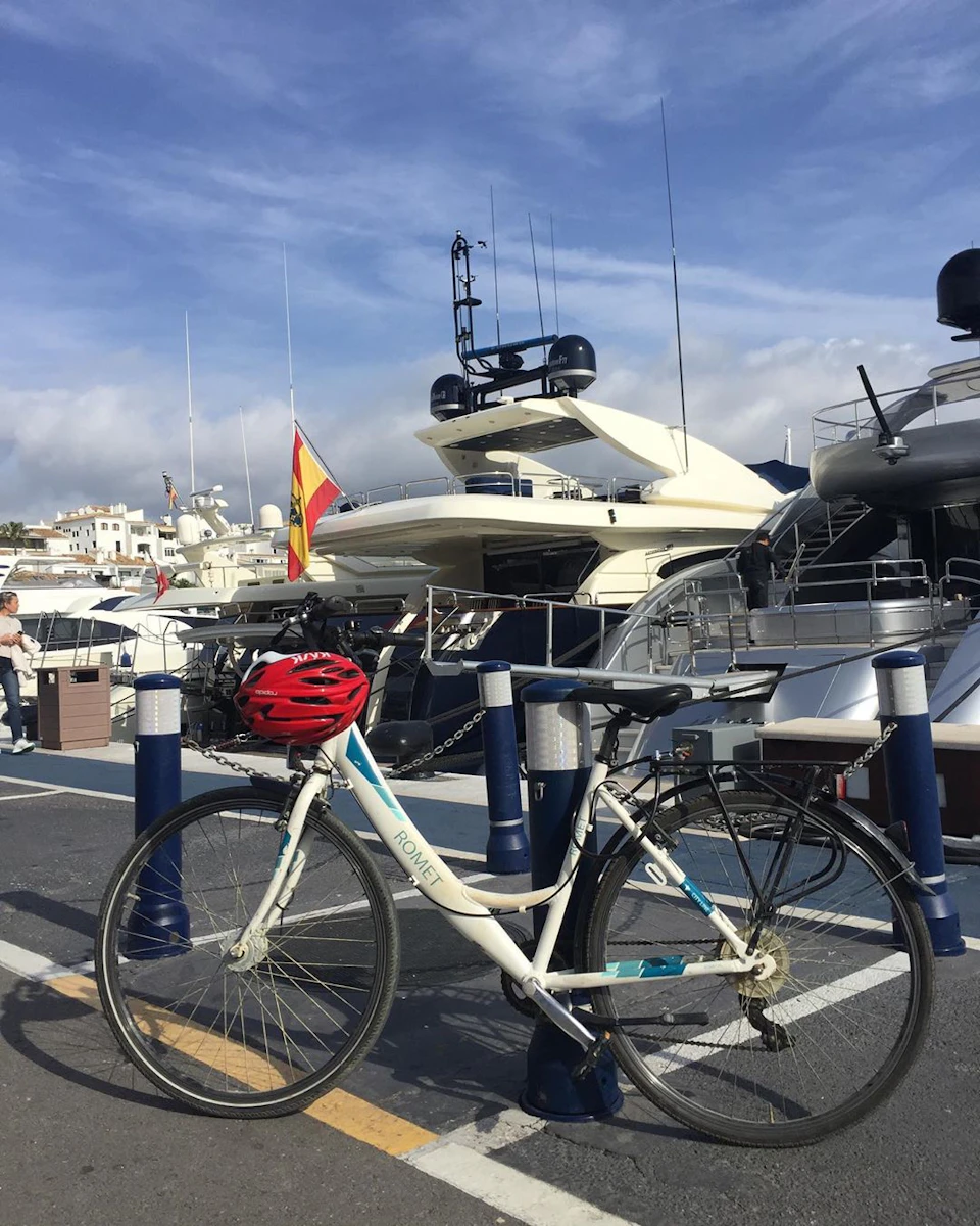 A bicycle with a red helmet parked near a row of yachts at a marina under a partly cloudy sky.