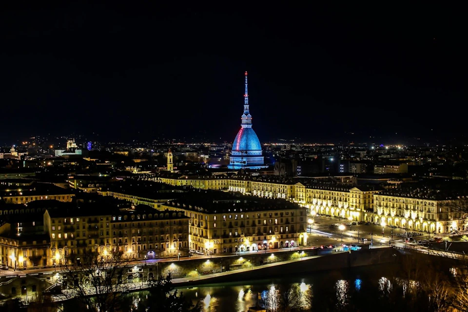 Night cityscape with a lit dome structure and surrounding illuminated buildings. A river flows in the foreground.