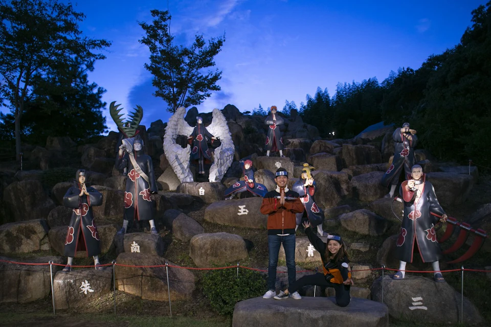 Two people pose in front of a display of anime character statues on a rocky background during twilight.
