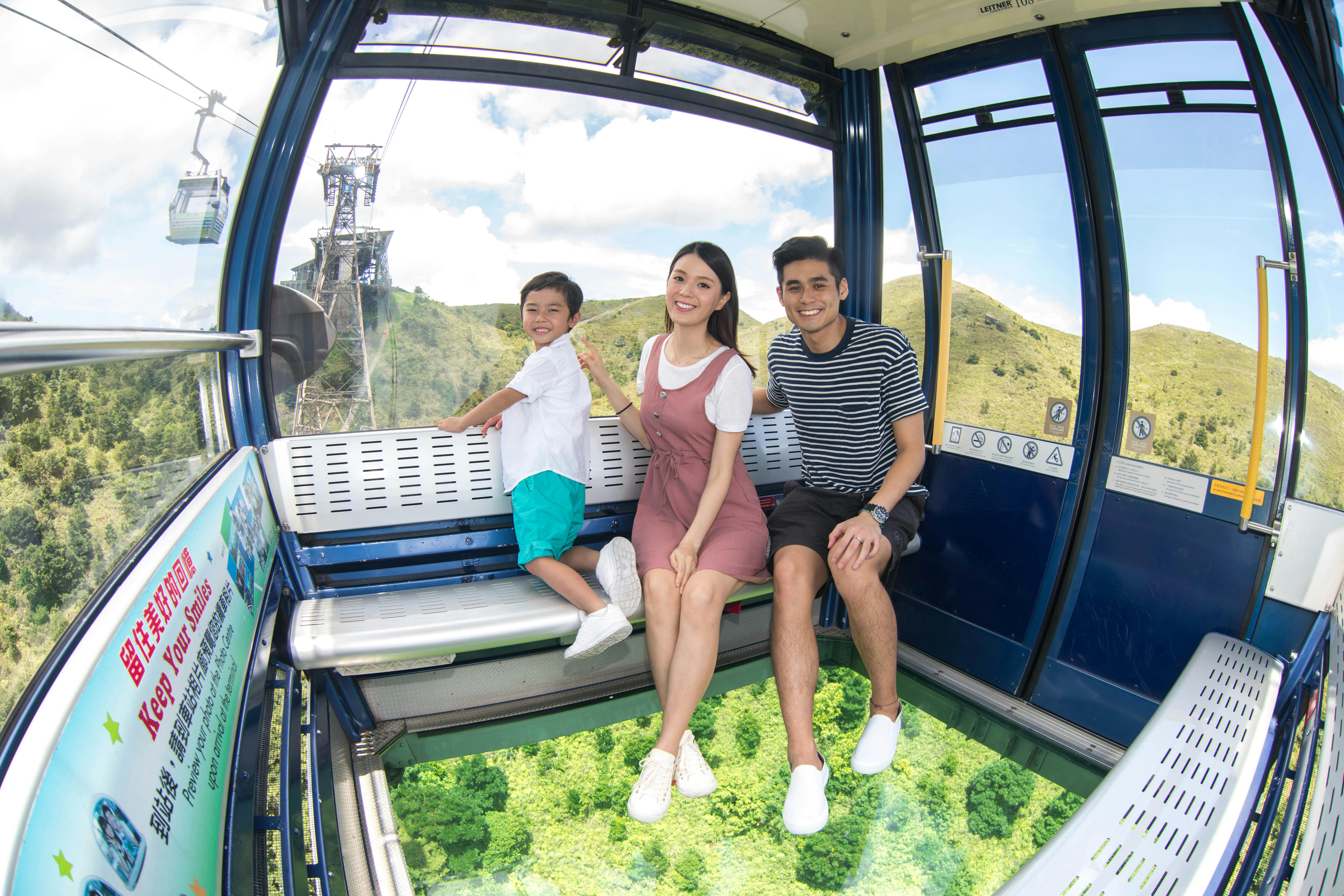 A family of three smiles while seated in a cable car with a glass floor, revealing green treetops below and hills in the background.