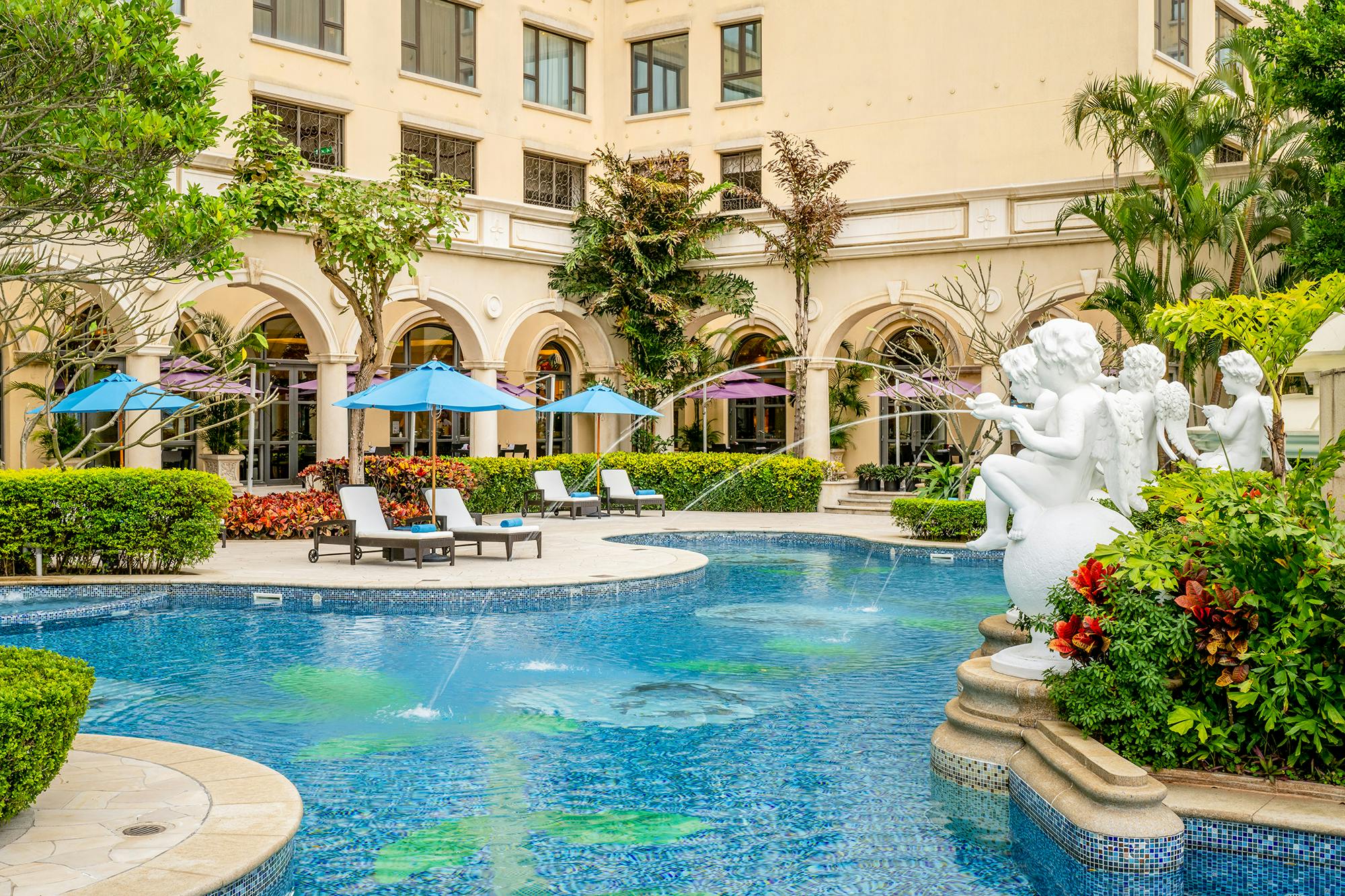 Outdoor pool area with lounge chairs, umbrellas, a fountain statue, and a building with arches and windows in the background.
