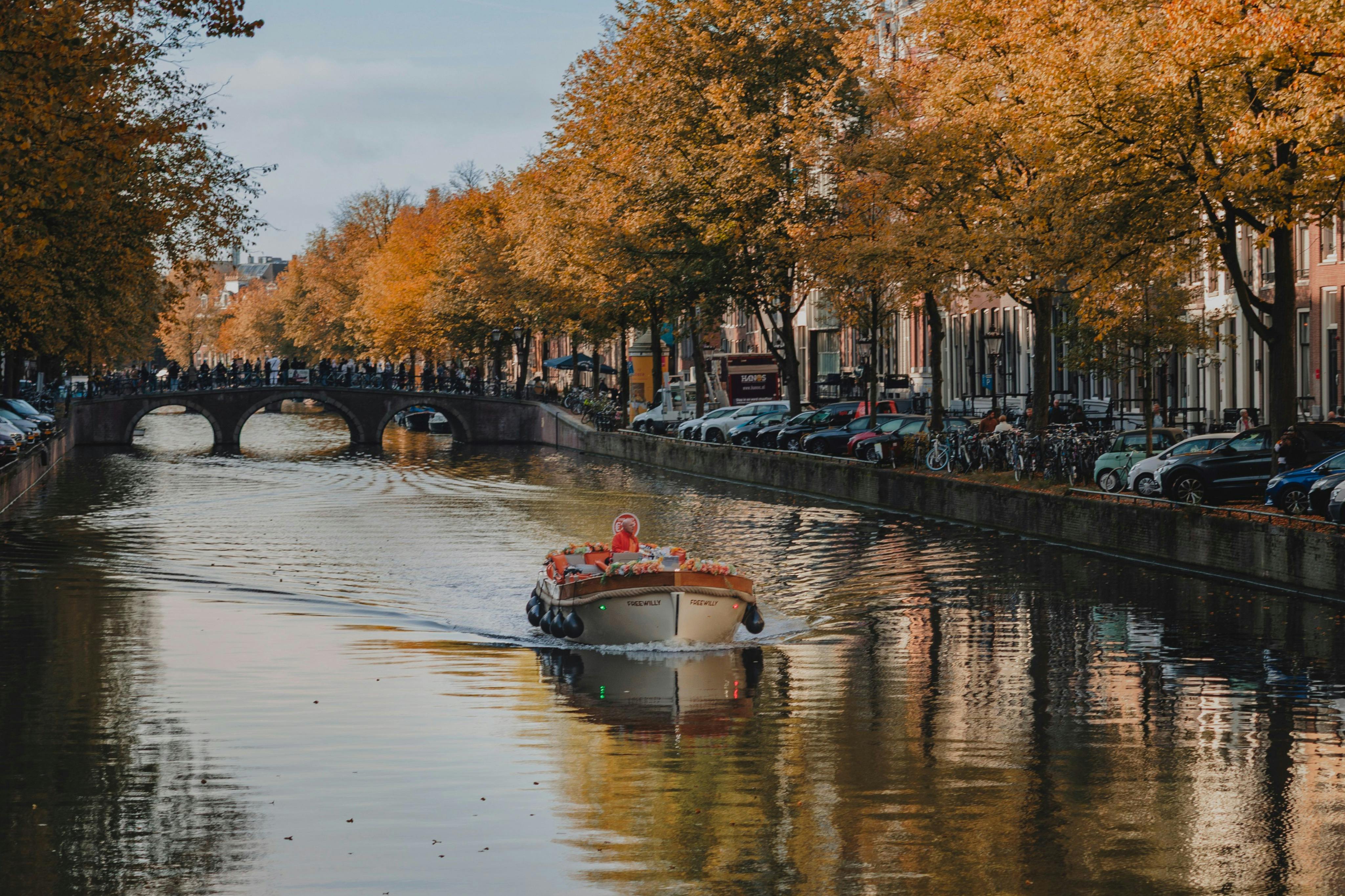 A boat cruises down a tree-lined canal with autumn foliage, parked cars, bicycles, and a bridge in the background.