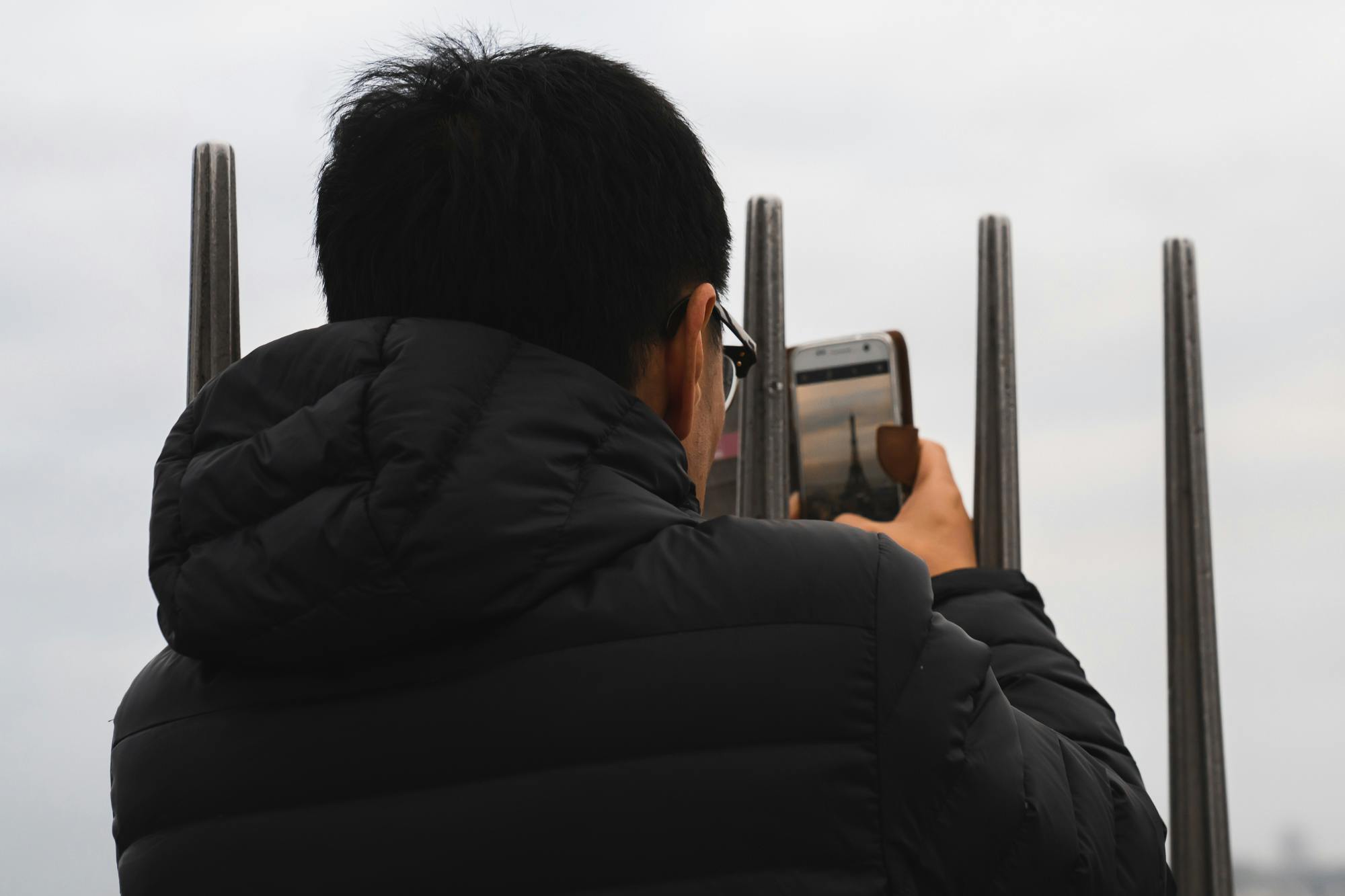 Person in a black jacket, taking a photo with a smartphone between metal bars, with a cloudy sky in the background.