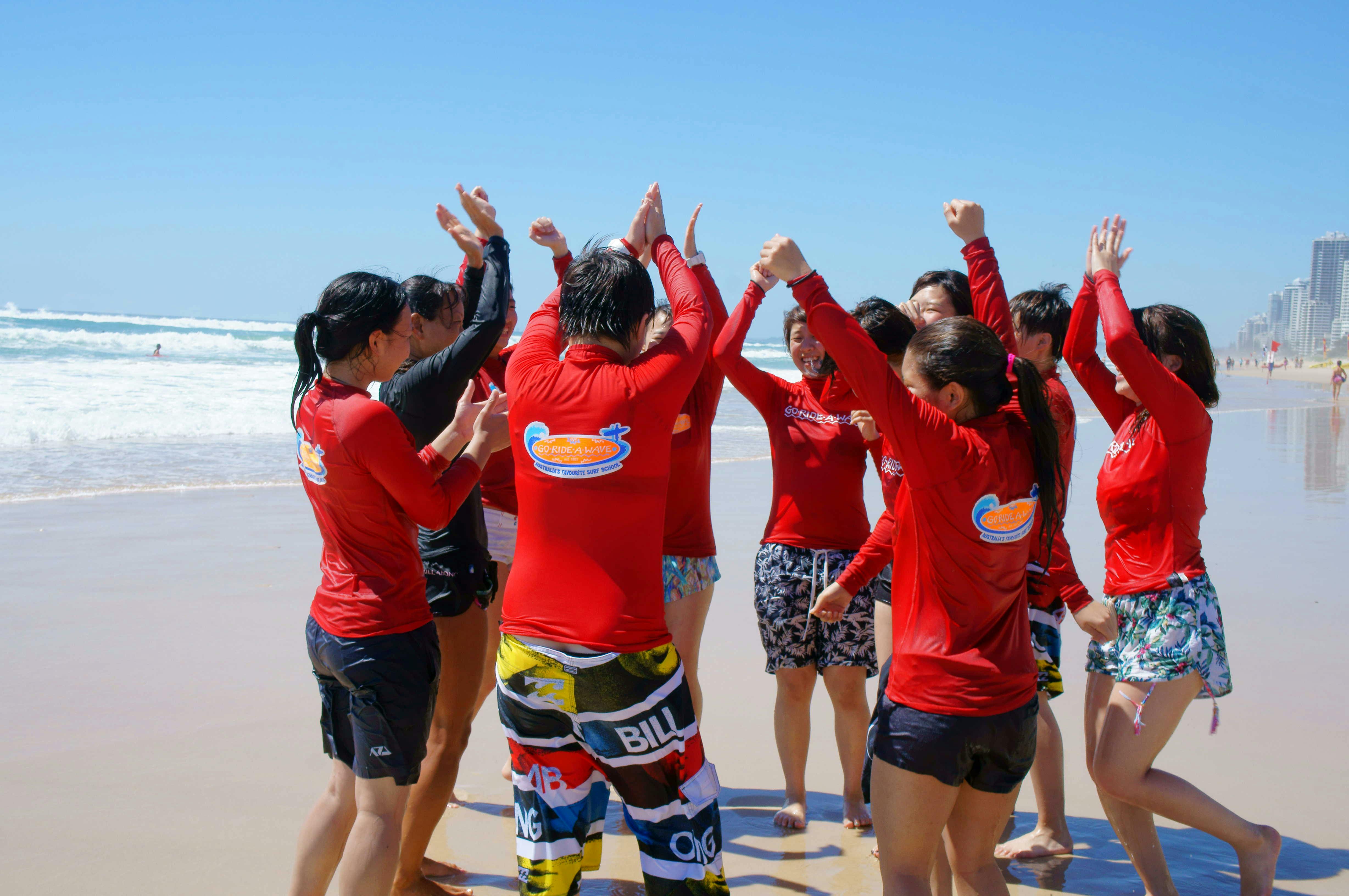 빨간 셔츠를 입고 해변에서 수영복을 입은 한 무리의 사람들이 맑고 푸른 하늘 아래에서 신나게 손을 들고 있습니다.