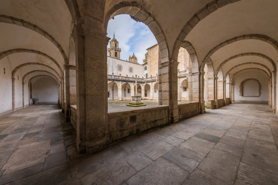 A stone courtyard surrounded by arched walkways, with towers and blue sky visible in the background.