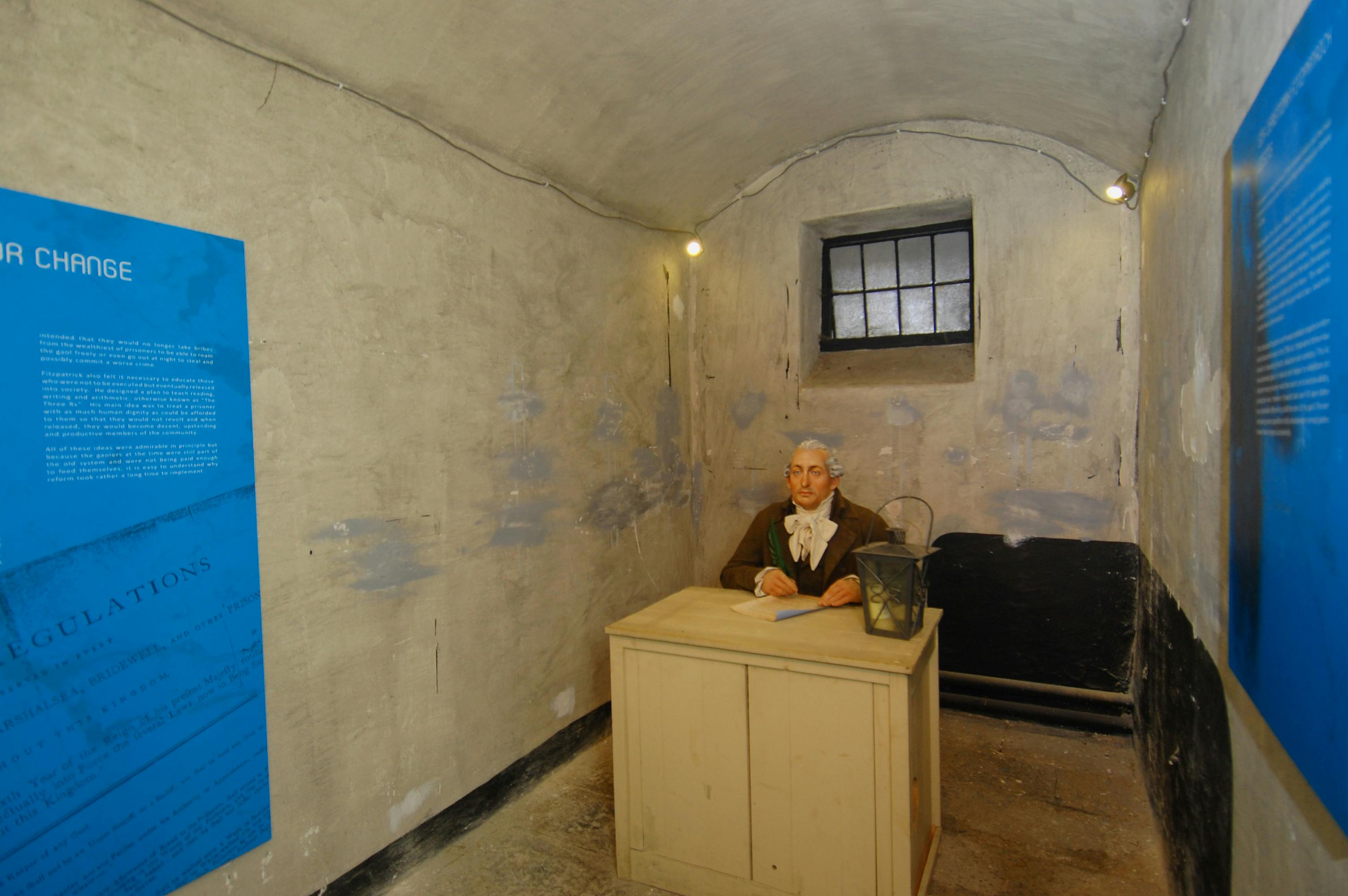 U dřevěného stolu s lucernou v malé, matně osvětlené betonové místnosti sedí vosková figurína oblečená v historickém oděvu.