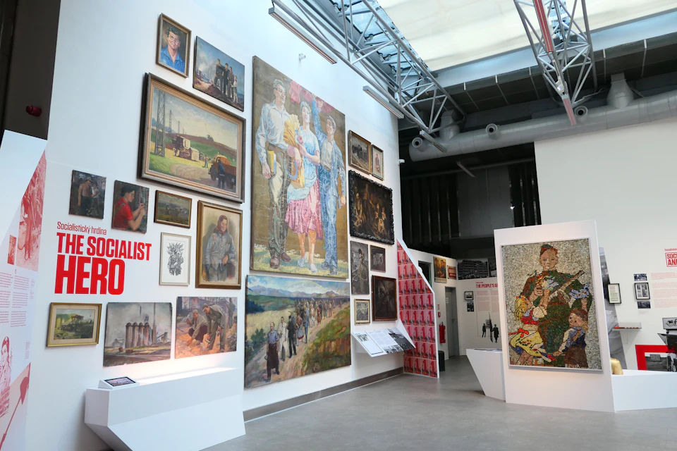 An art gallery wall featuring paintings and photographs, predominantly showcasing people and landscapes, under industrial lighting.
