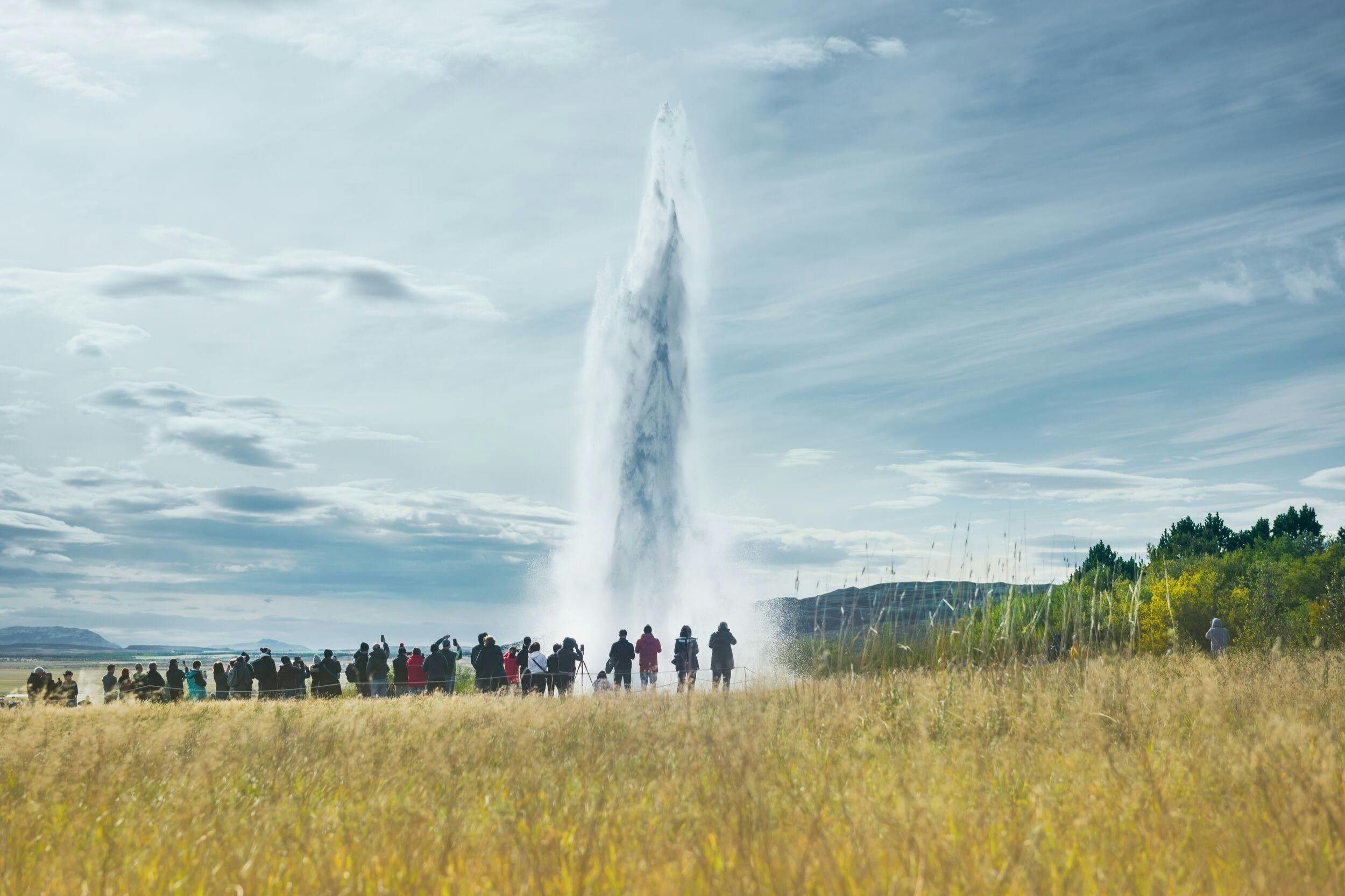 A group of people observing a tall geyser eruption in a grassy landscape under a partly cloudy sky.