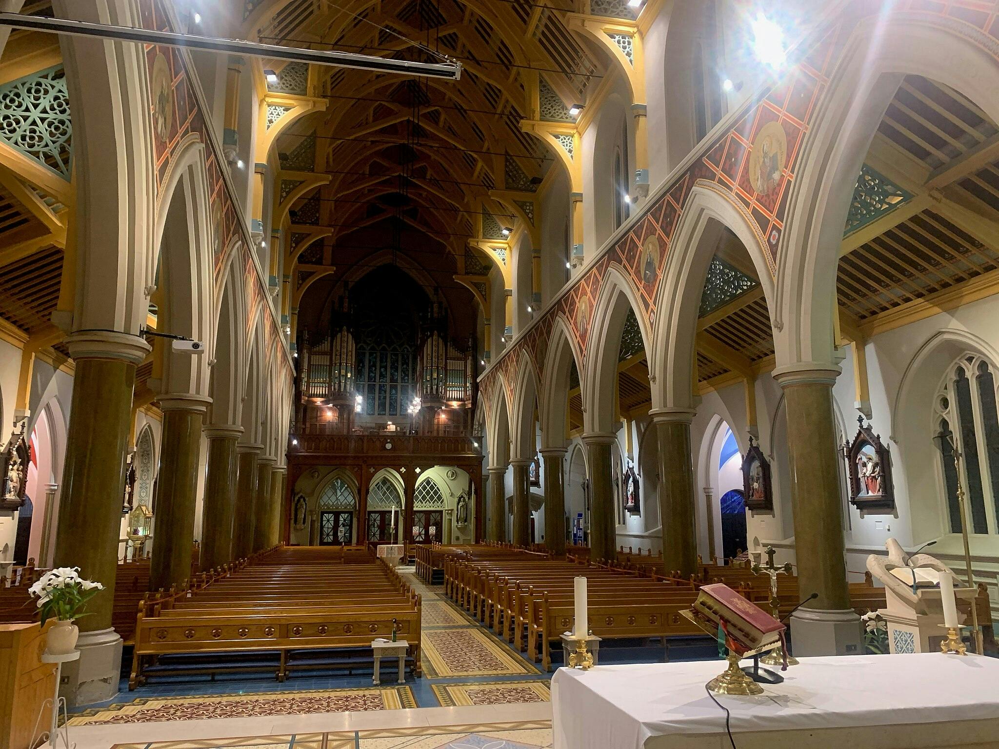 Interior of a church with wooden pews, ornate columns, and vaulted ceilings. An altar with candles and a book is in the foreground.