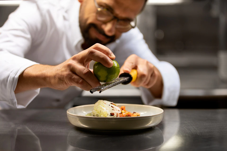 A chef zesting a lime onto a plated dish, focusing on the food preparation in a professional kitchen.