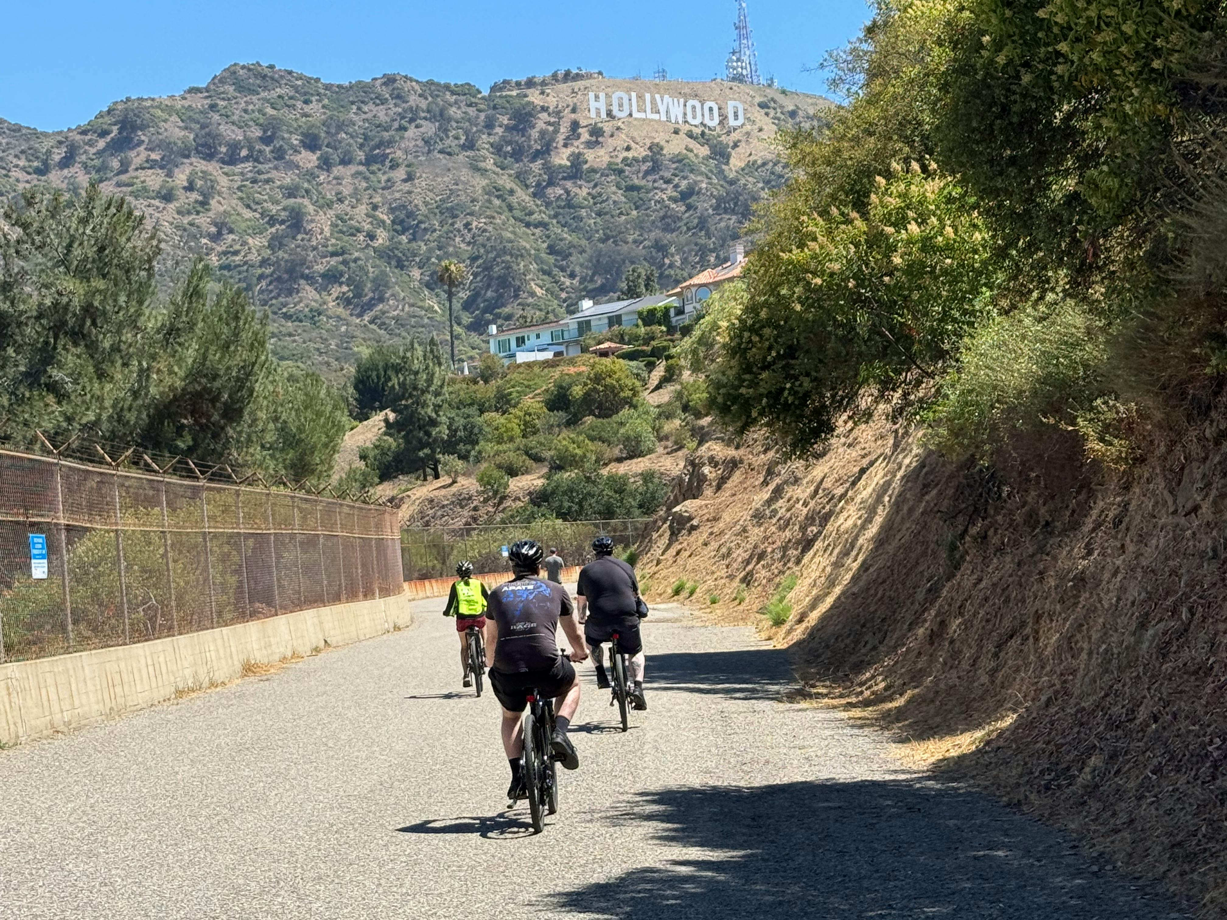 Three cyclists ride on a paved road with the Hollywood sign visible on a shrub-covered hill in the background.