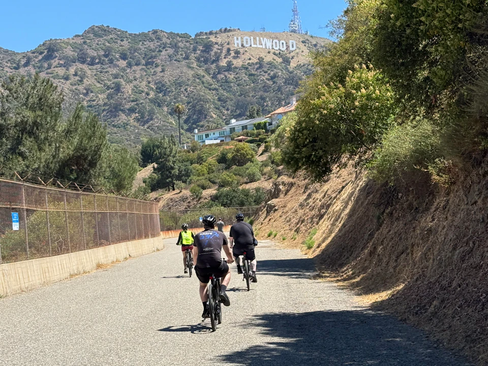 Three cyclists ride on a paved road with the Hollywood sign visible on a shrub-covered hill in the background.