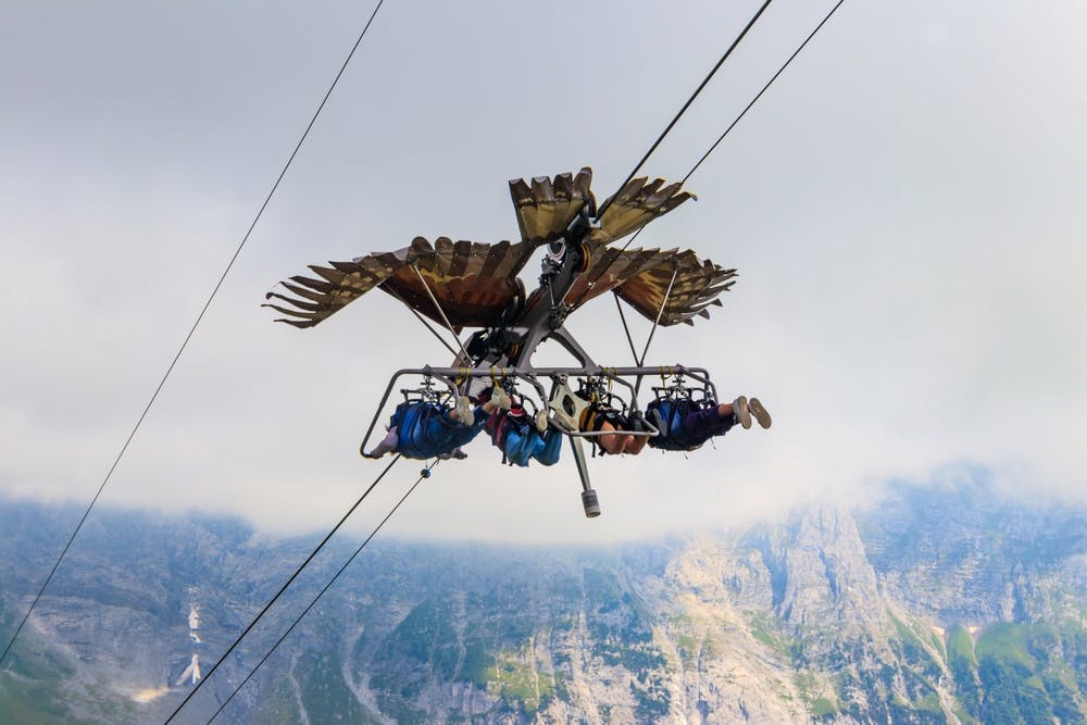 Lidé v postrojích visí na zipline ve tvaru ptáka na hornatém pozadí.