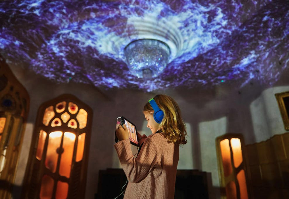 A child wearing headphones holds a tablet in a room with ornate decor and a ceiling lit by colorful projections.