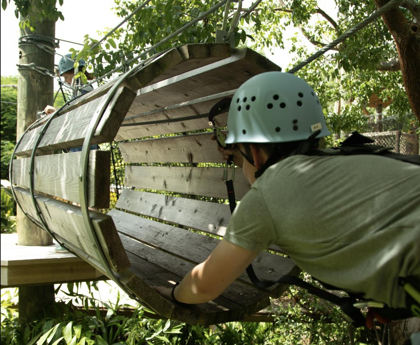 ヘルメットをかぶった人が木製のトンネルに入り、緑豊かな屋外に吊るされています。