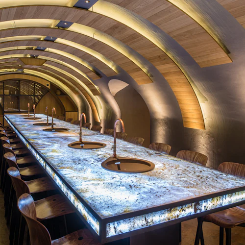 En modern bar med ett välvt trätak, bänkskiva i marmor och trästolar, upplyst av varm, integrerad belysning.