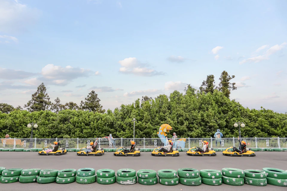 People driving small bumper cars on a track surrounded by green tires, with trees and a clear sky in the background.