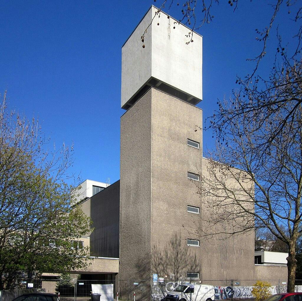 Tall concrete tower with rectangular windows, topped by a large cubic structure, surrounded by bare trees and a blue sky.