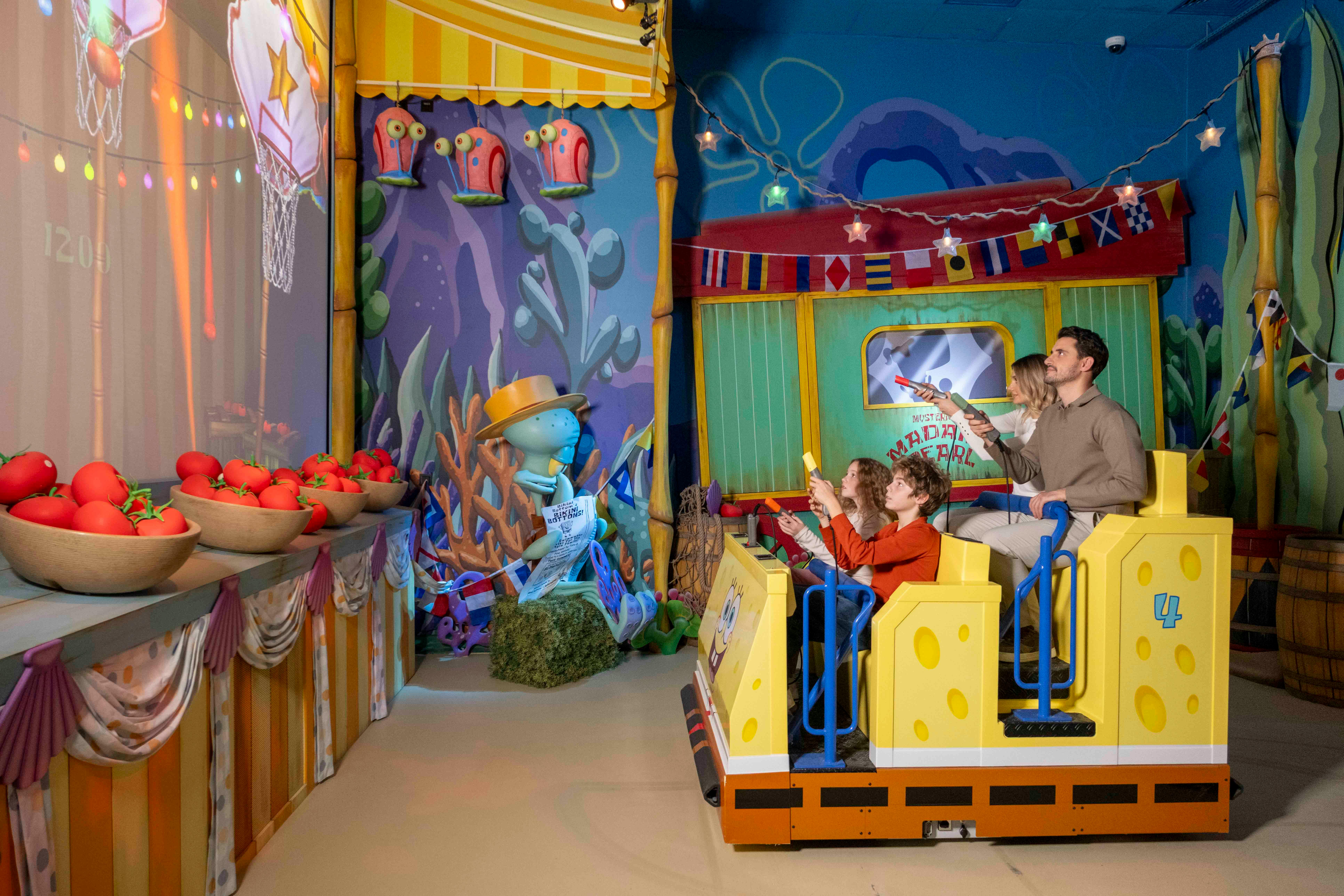 Mensen zitten in een geel voertuig in een kleurrijke indoorattractie met cartoon-thema en houden rekwisieten in 3D-stijl vast.