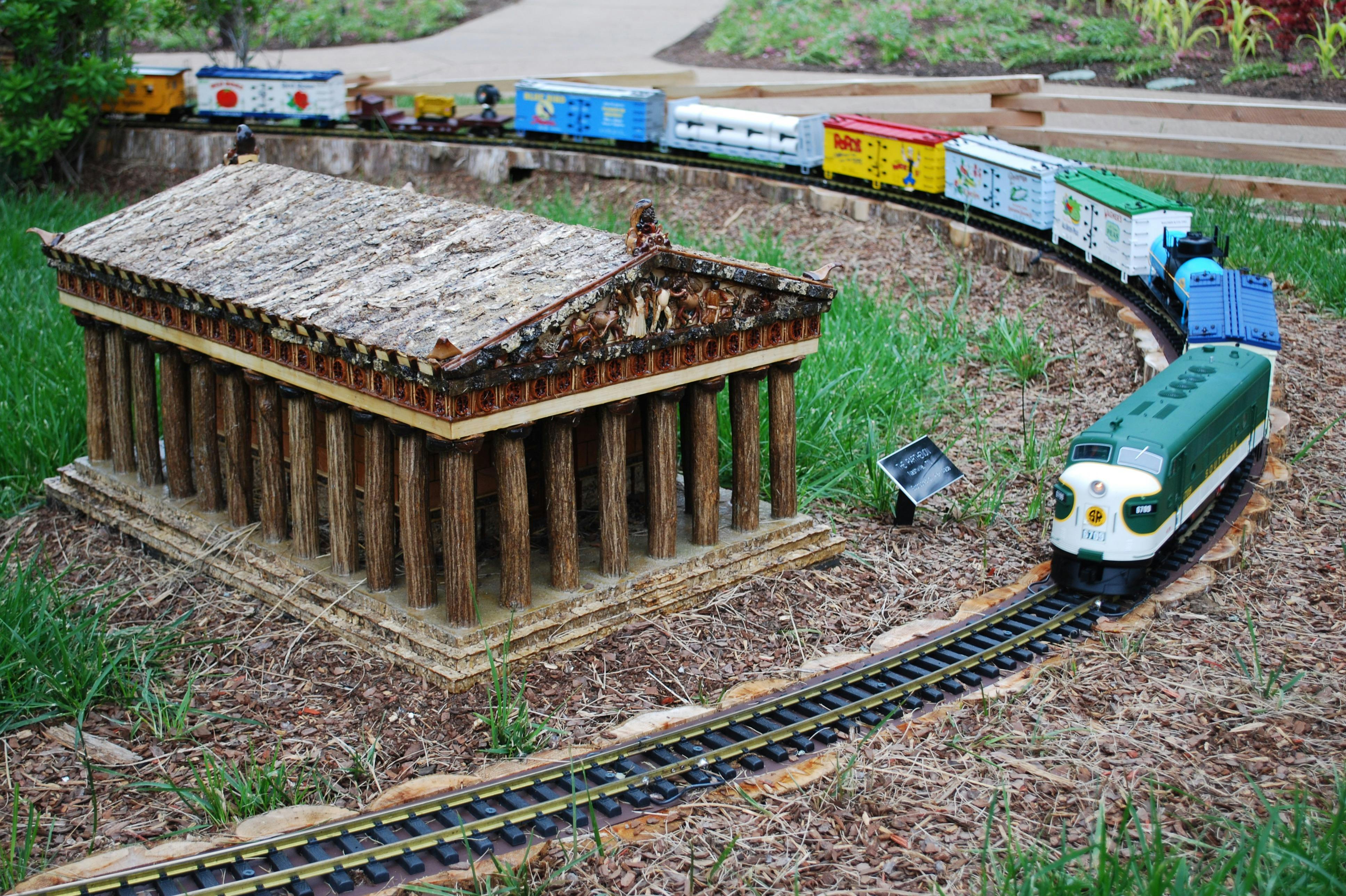 Miniaturní model řeckého chrámu s malými vlaky na kolejích, které jej obklopují ve venkovním prostředí.