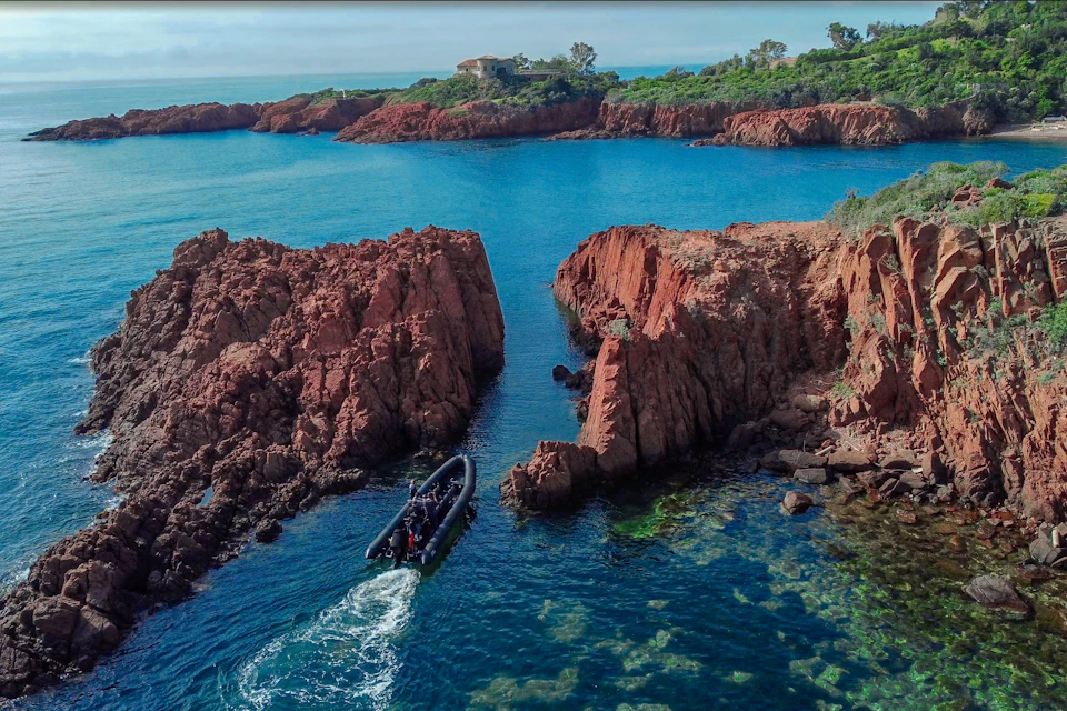 A small boat navigating between red rock formations on a blue sea, with a distant shoreline and greenery in the background.