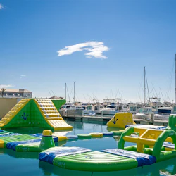 Inflatable water park with slides and obstacles floats on water, with a marina full of boats in the background under a clear sky.