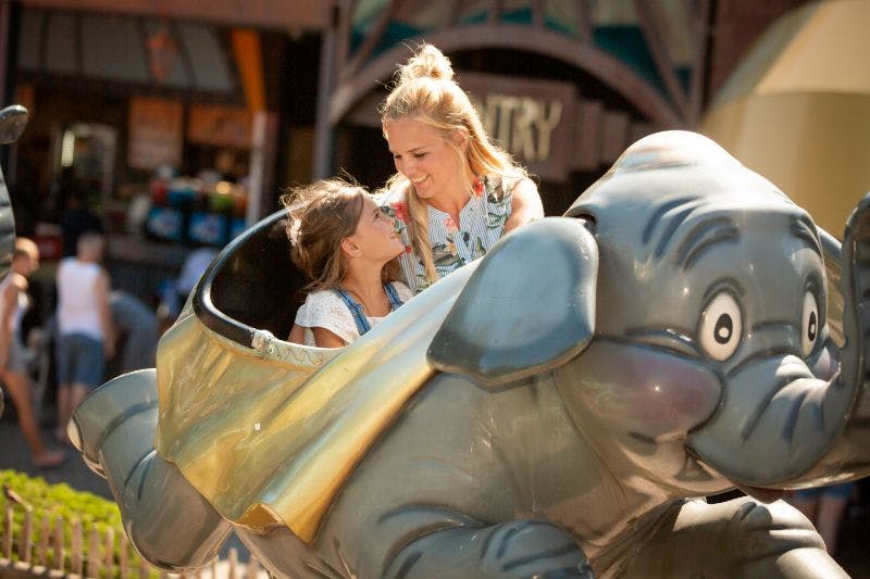 A woman and a young girl smiling at each other while riding an elephant-themed amusement park ride.