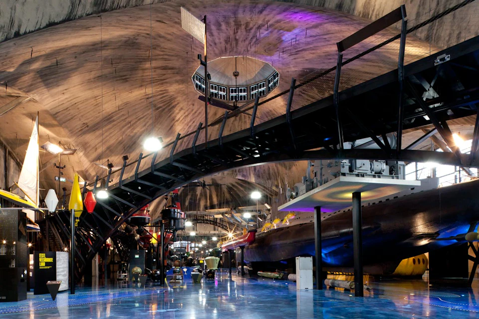 Interior of a dimly lit museum with industrial design, featuring a submarine exhibit, overhead walkways, and vibrant lighting.