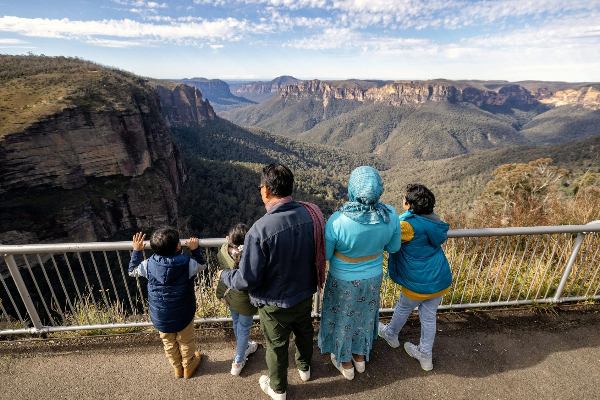 A family of five stands at a railing overlooking a vast canyon landscape with cliffs and forested valleys under a partly cloudy sky.