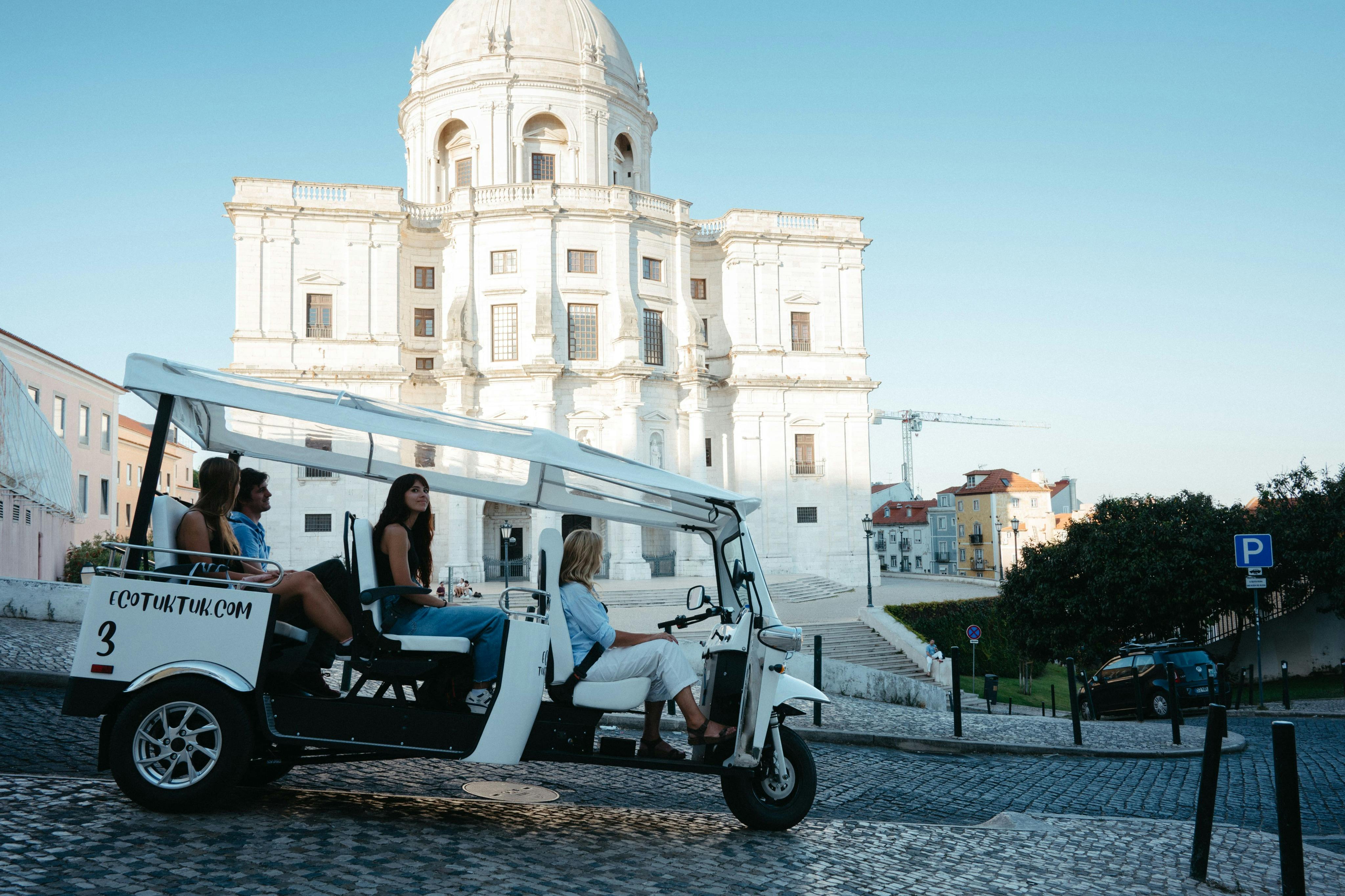 People sitting in a white tuk-tuk vehicle parked on a cobblestone street in front of a white dome building on a clear day.