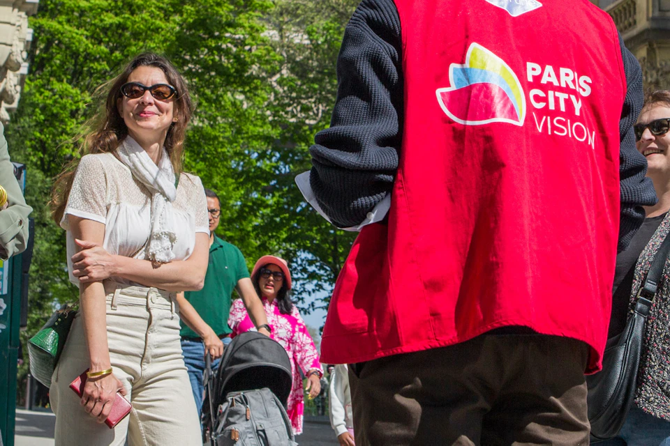 A guide wearing a red vest labeled 'Paris City Vision' talking to a group of tourists near the Eiffel Tower on a sunny day.