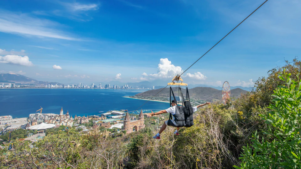 A person gliding on a zipline over a scenic coastal area with buildings, water, and hills under a blue sky.