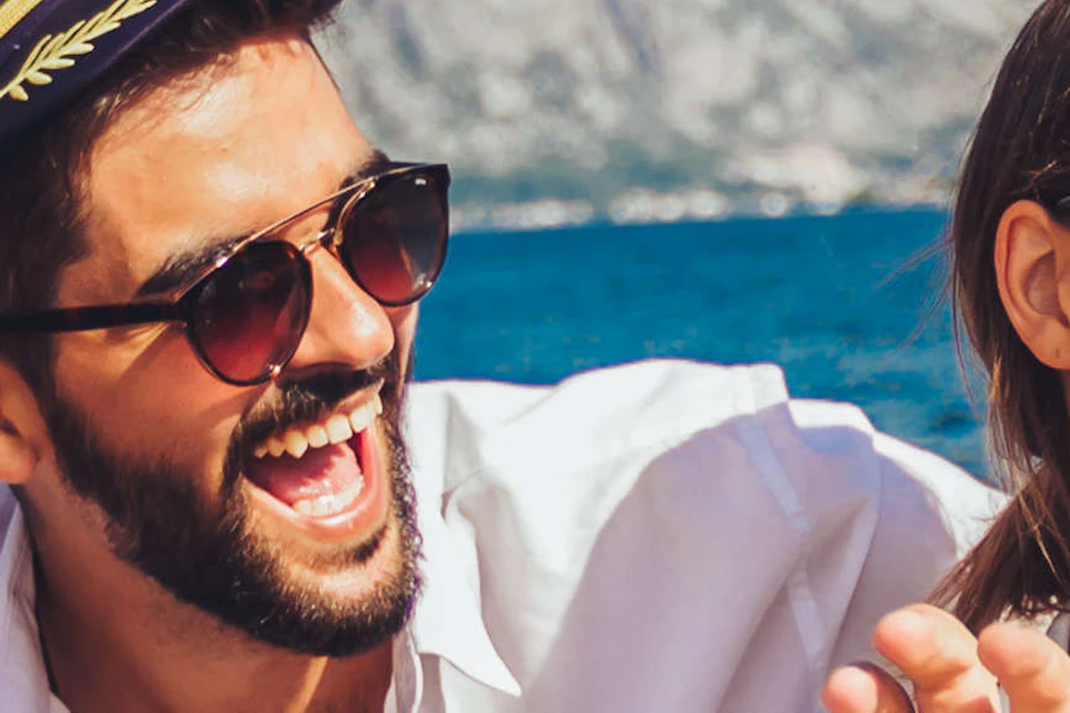 Four friends laughing and enjoying drinks on a boat, with mountains and blue sky in the background.